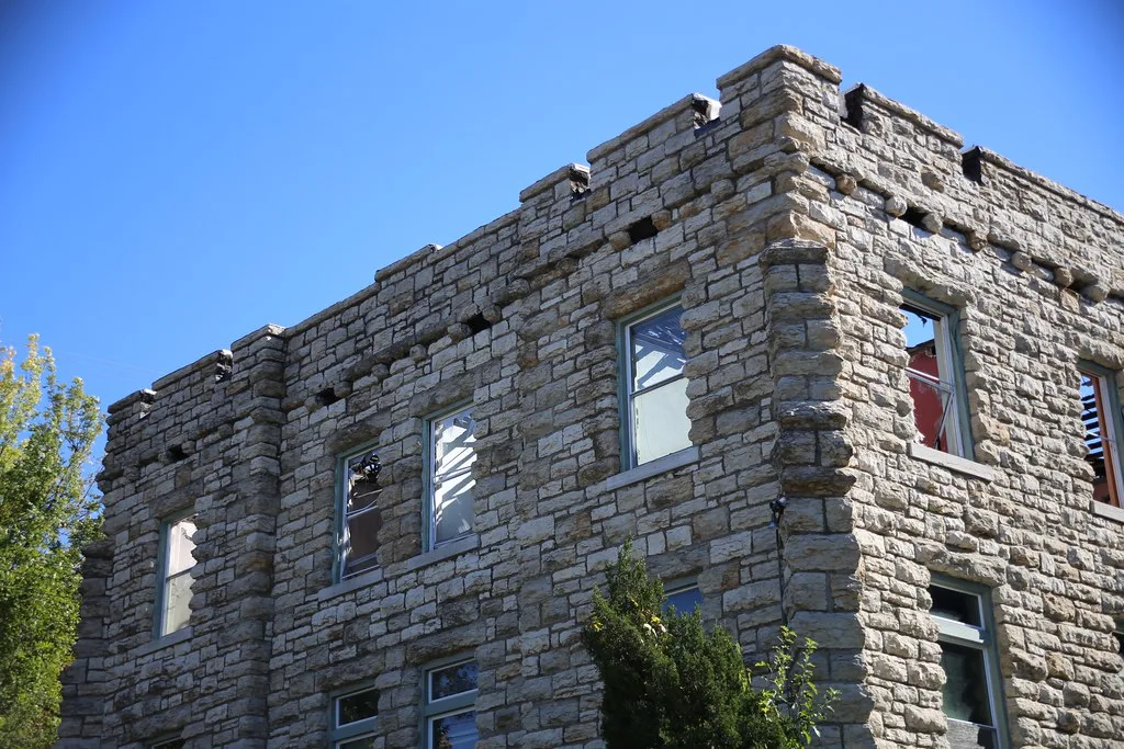 Corner of an abandoned pale stone building without a roof, with broken windows and remaining roof framing against blue sky.