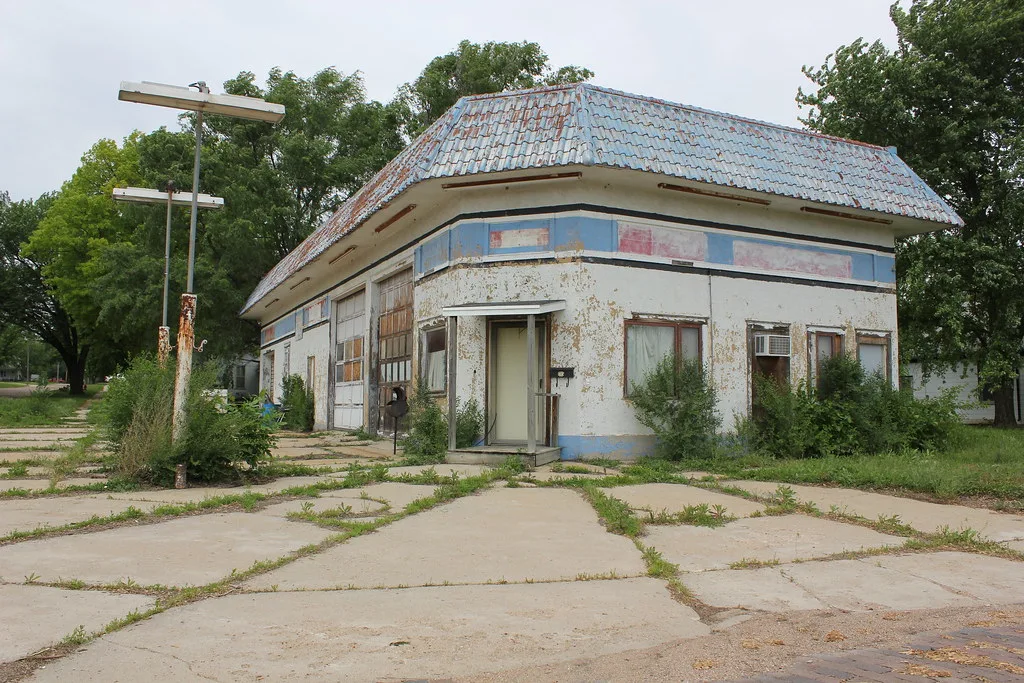 Former garage with a rusty blue roof, peeling paint and tall weeds in front of its glazed doors.