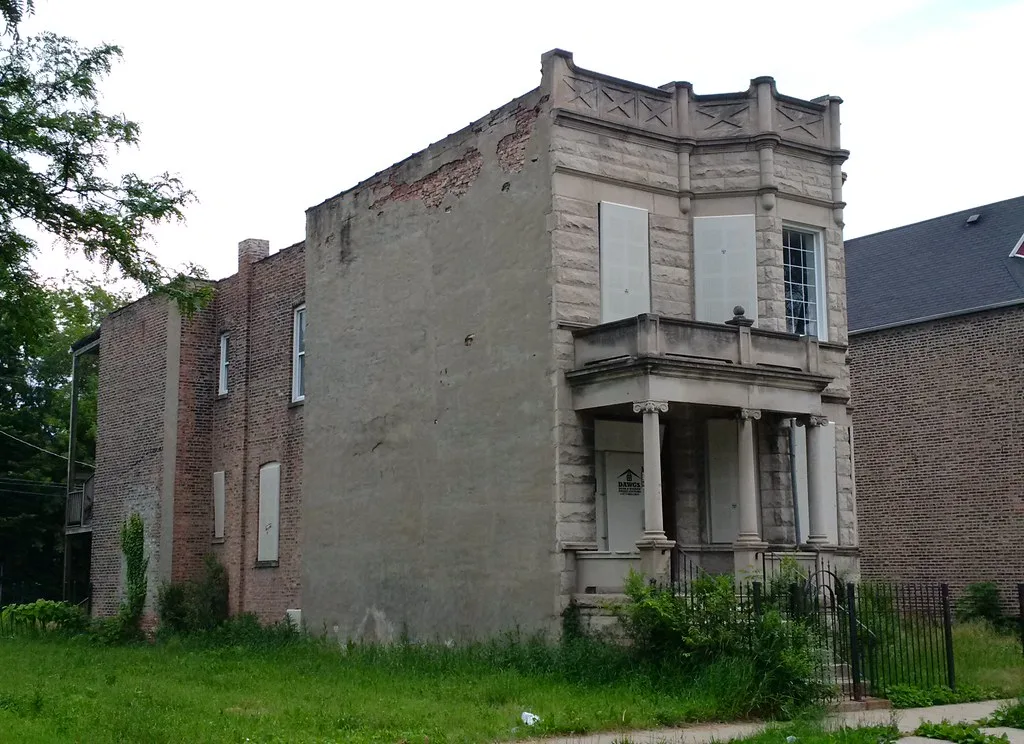 Abandoned house with a columned porch, sealed openings and missing render above an overgrown garden.