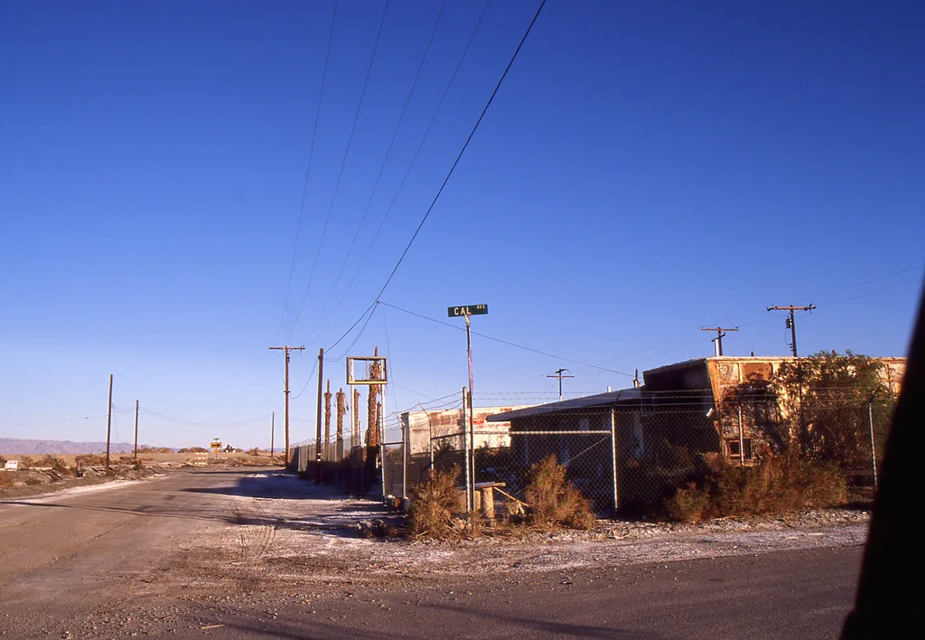 Abandoned buildings beside a desert street behind a scrub-covered fence.