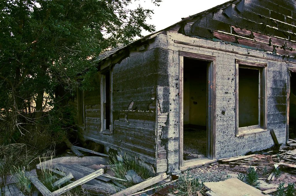 Small abandoned house with empty openings, torn gray cladding and fallen boards in front of the facade.