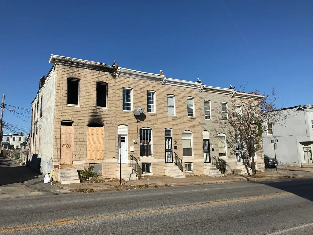 Brick house with blackened boarded windows beside intact houses.