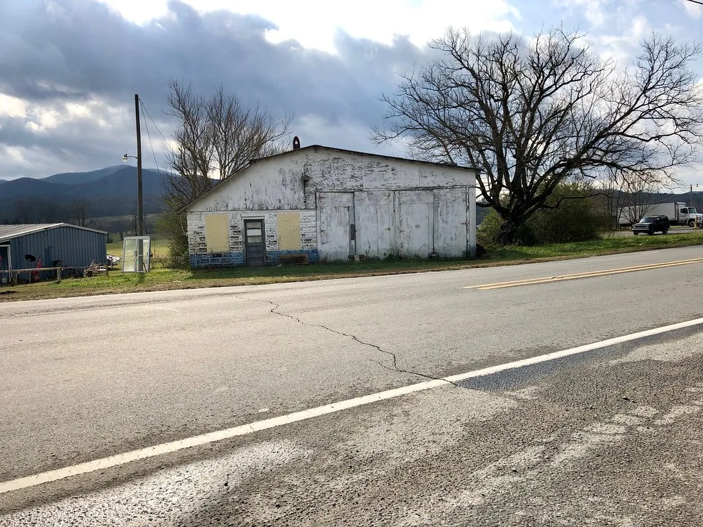 Old low white building with boarded windows beside a road.
