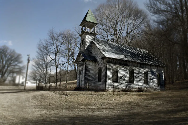 Small weathered wooden church with boarded windows and an open bell tower before leafless trees.