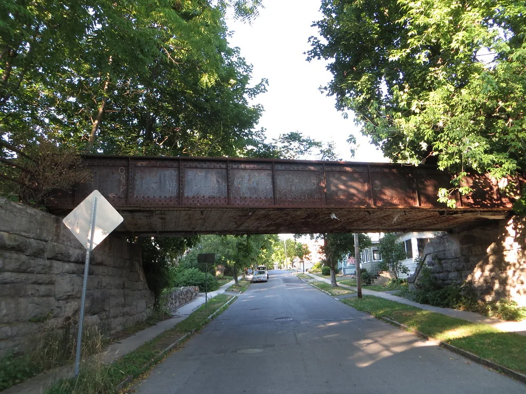 Ancien pont métallique rouillé au-dessus d’une rue bordée de maisons.