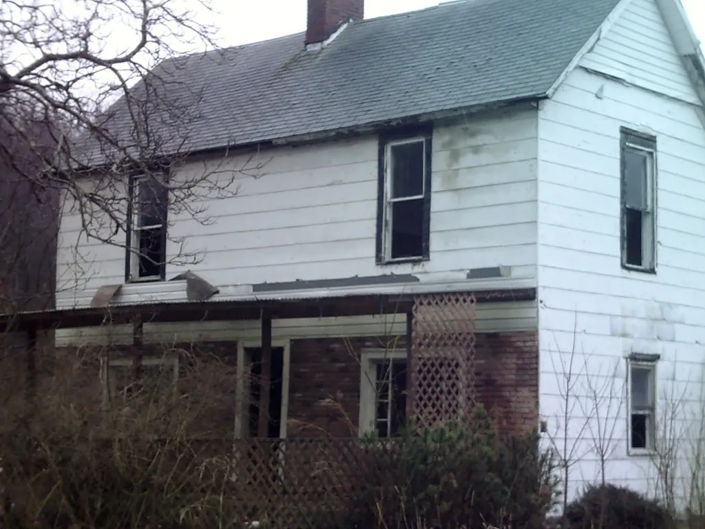 Neglected white house with broken windows and an overgrown porch.