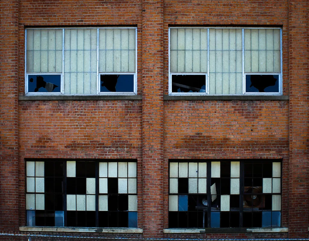 Brick facade of an abandoned factory with four large windows containing many broken panes.