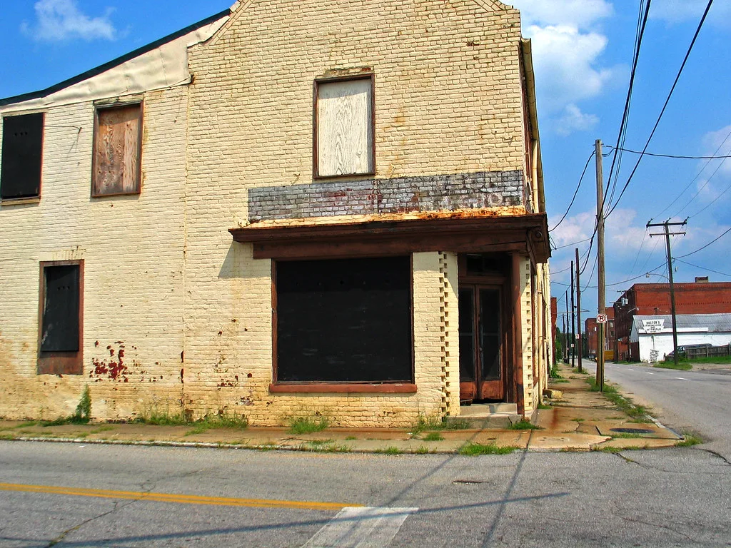 Old yellow brick shop with boarded openings on a street corner.