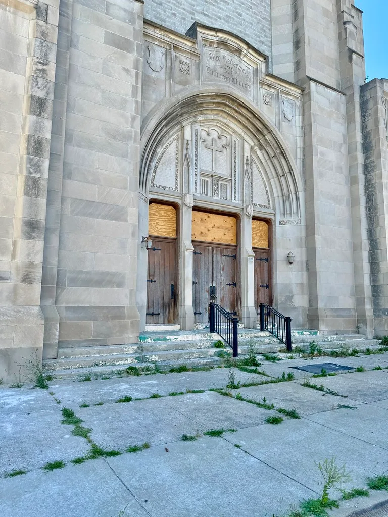 Entrance of a neglected church with wooden doors and boarded transoms above damaged steps.