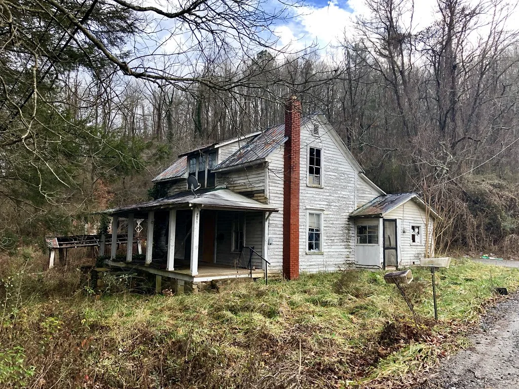 Maison abandonnée blanche à cheminée de briques et porche délabré, aux fenêtres cassées devant une forêt.