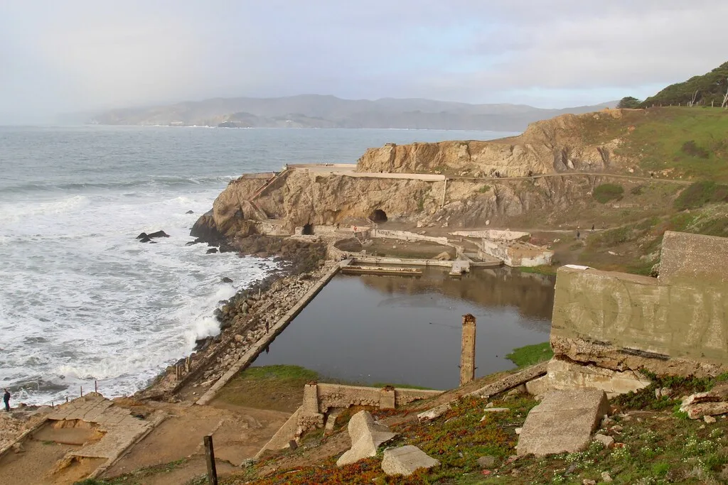 Ruins of coastal pools with broken concrete walls and slabs, below a cliff facing ocean waves.