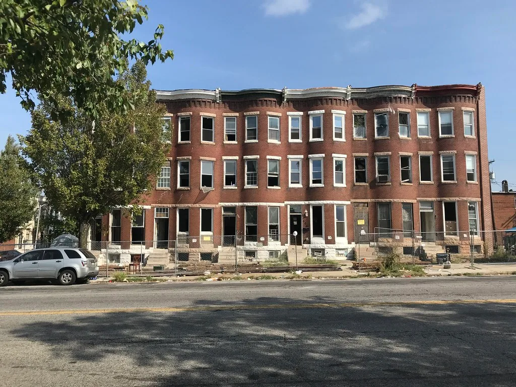 Row of brick houses with broken windows behind a construction fence.