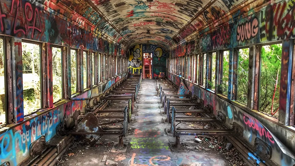 Abandoned railway carriage interior covered in graffiti, with stripped seats and empty windows.