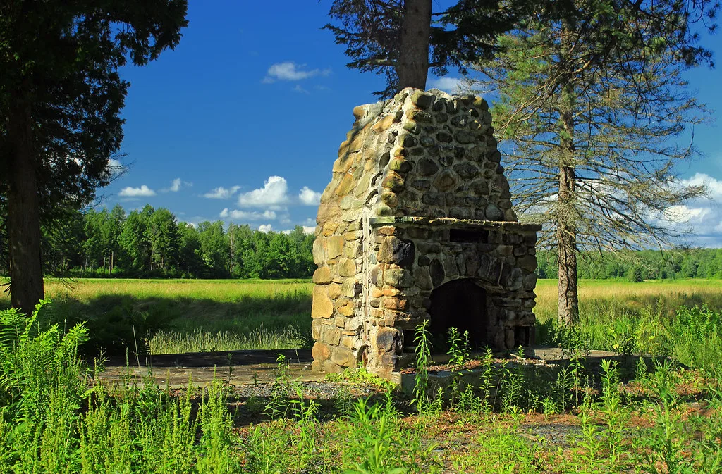 Cheminée abandonnée en pierres arrondies seule sur une dalle de fondation couverte de plantes, au bord d’une prairie.
