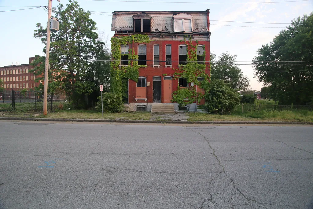 Abandoned red-brick house with broken windows and a deteriorated mansard roof covered in ivy.