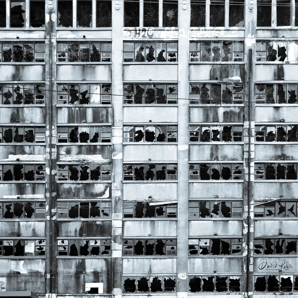 Black-and-white abandoned factory facade with rows of shattered windows separated by concrete pillars.