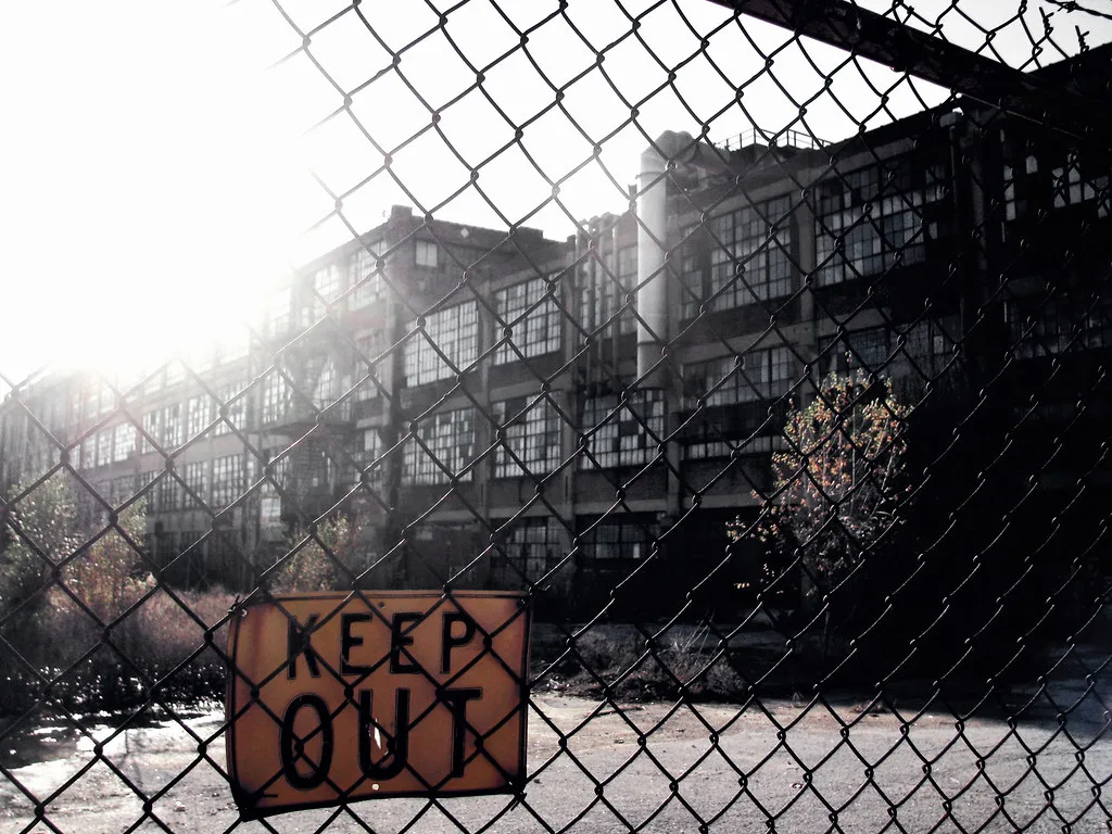 Abandoned factory behind a wire fence bearing a keep-out sign.