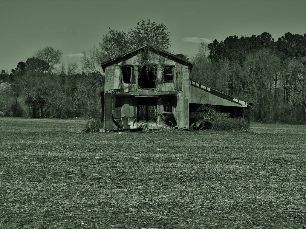 Old two-story rural building with gaping windows and torn siding beside a field.
