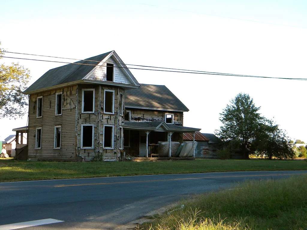 Old house with empty windows and torn siding beside a road.