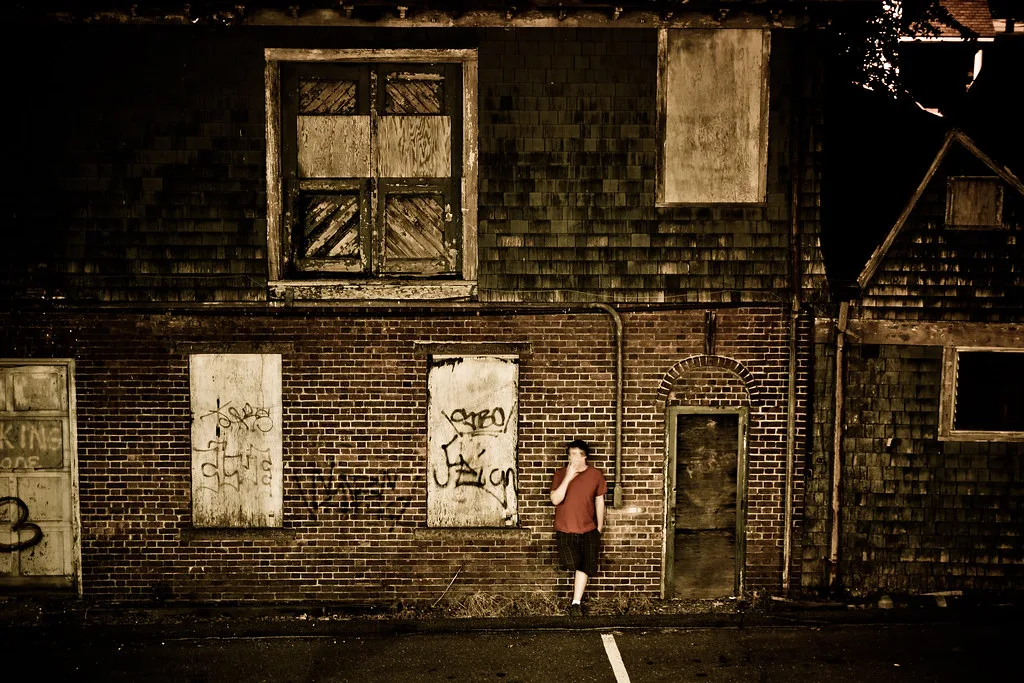 Abandoned brick and shingle facade with boarded openings and a person standing against the wall.