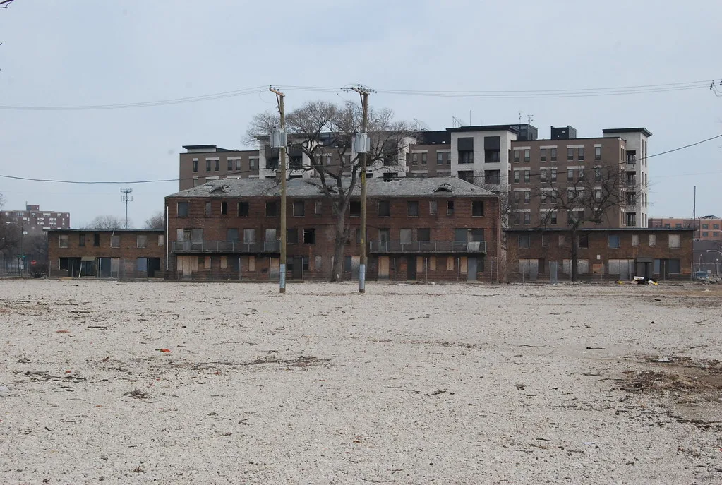 Brick buildings with boarded openings in front of a newer apartment building.
