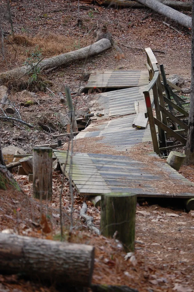 Ruined wooden footbridge with a sagging deck and missing boards in woodland.