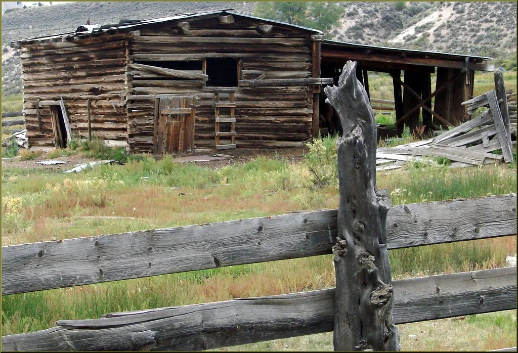 Dilapidated old log barn behind a wooden fence.