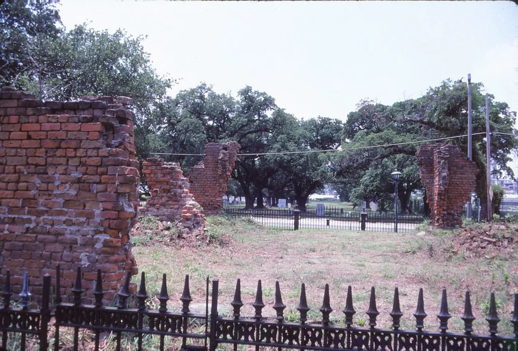 Ruins of roofless red brick walls, with rubble and grass behind a black fence among trees.