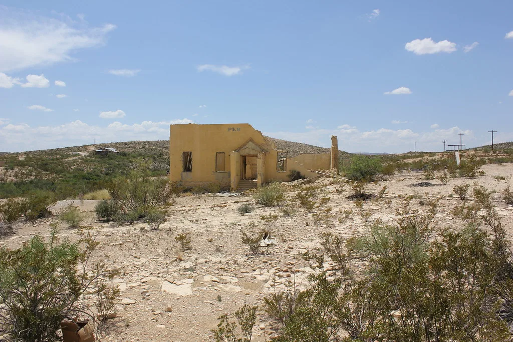 Roofless yellow building ruin with a pedimented entrance and isolated chimney on desert ground.