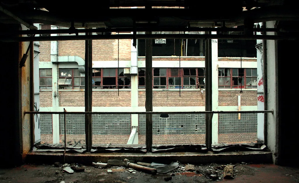 Grande baie métallique vide ouvrant sur une façade d’usine aux vitrages brisés.