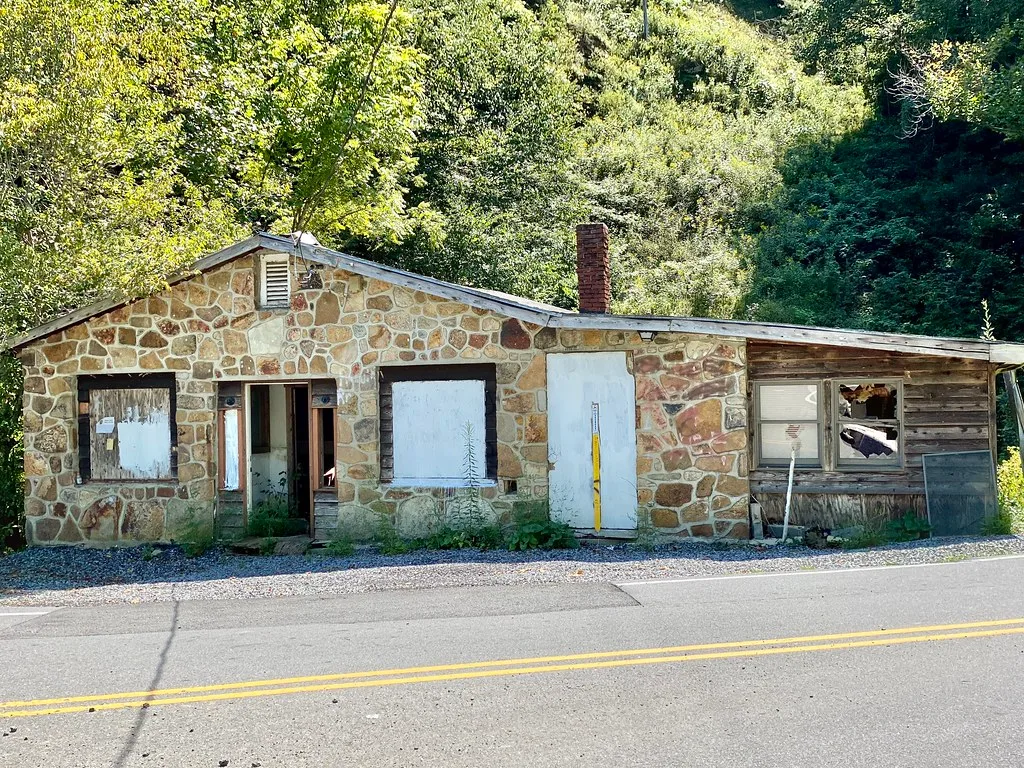 Petit bâtiment abandonné à parement de pierre et annexe en bois, fenêtres condamnées ou cassées en bord de route.