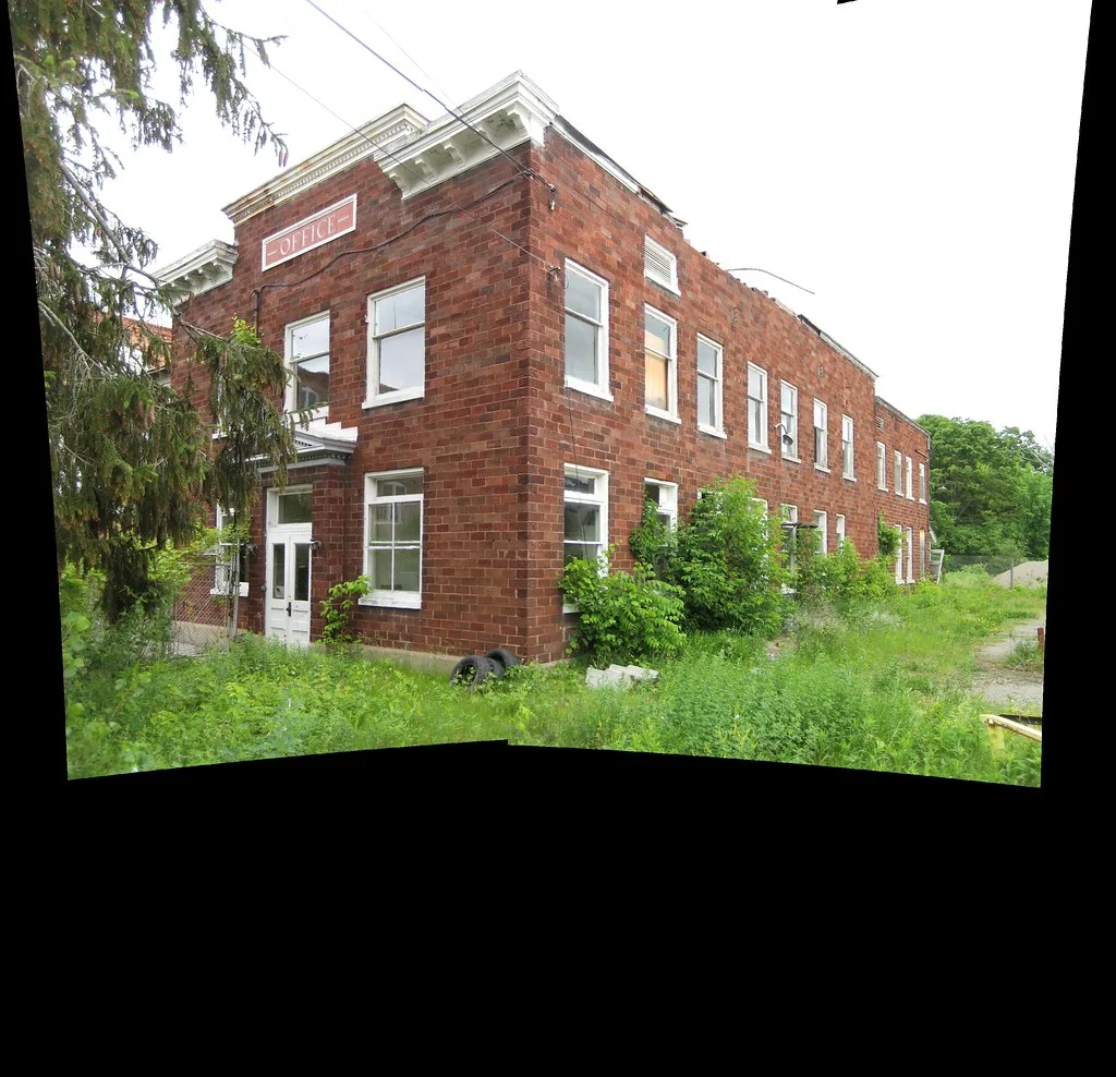Old red brick building overgrown with vegetation, in a stitched panorama.