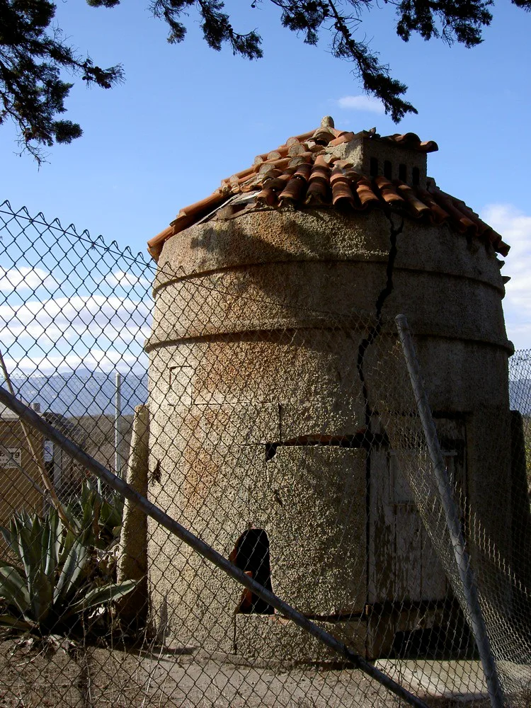 Neglected small cylindrical tower with a wide vertical crack beneath a damaged tiled roof.