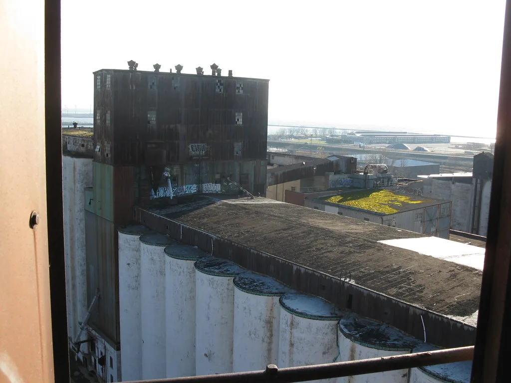Anciens silos et tour industrielle taguée vus à travers une fenêtre.