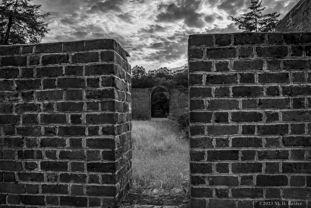 Roofless brick building ruins with a gap between two walls and an arch beyond a grassy interior.