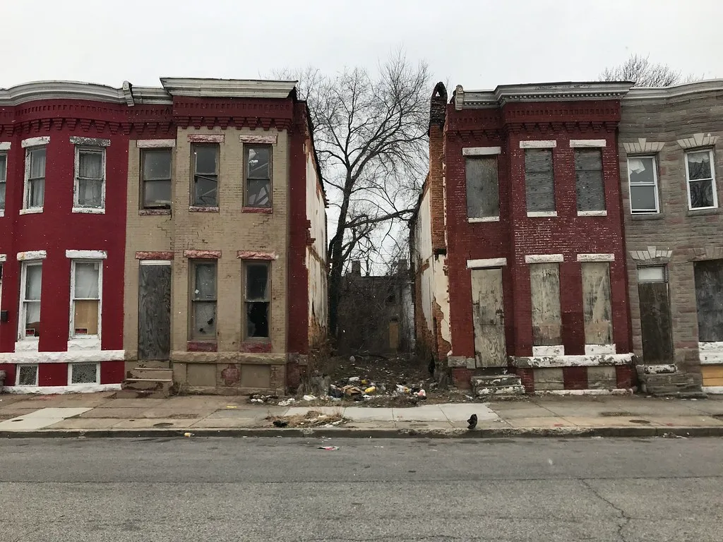 Abandoned red- and yellow-brick houses with boarded windows separated by a debris-strewn plot.