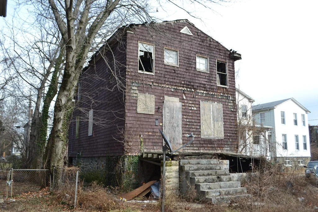 Maison abandonnée à bardage brun et fenêtres cassées, avec porte condamnée au-dessus d’un porche affaissé.