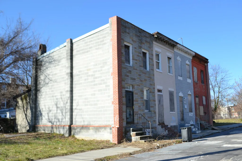 Row of small grey and red houses with boarded windows and weeds at the foot of the steps.