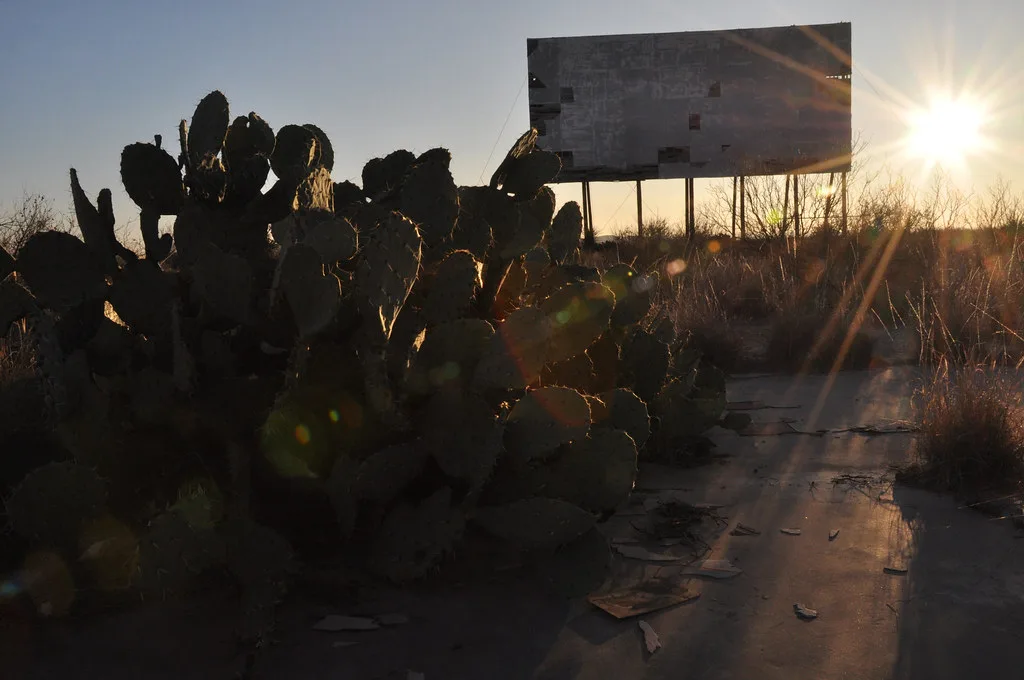 Écran abandonné de cinéma de plein air aux panneaux troués derrière des cactus et un sol jonché de débris au soleil couchant.