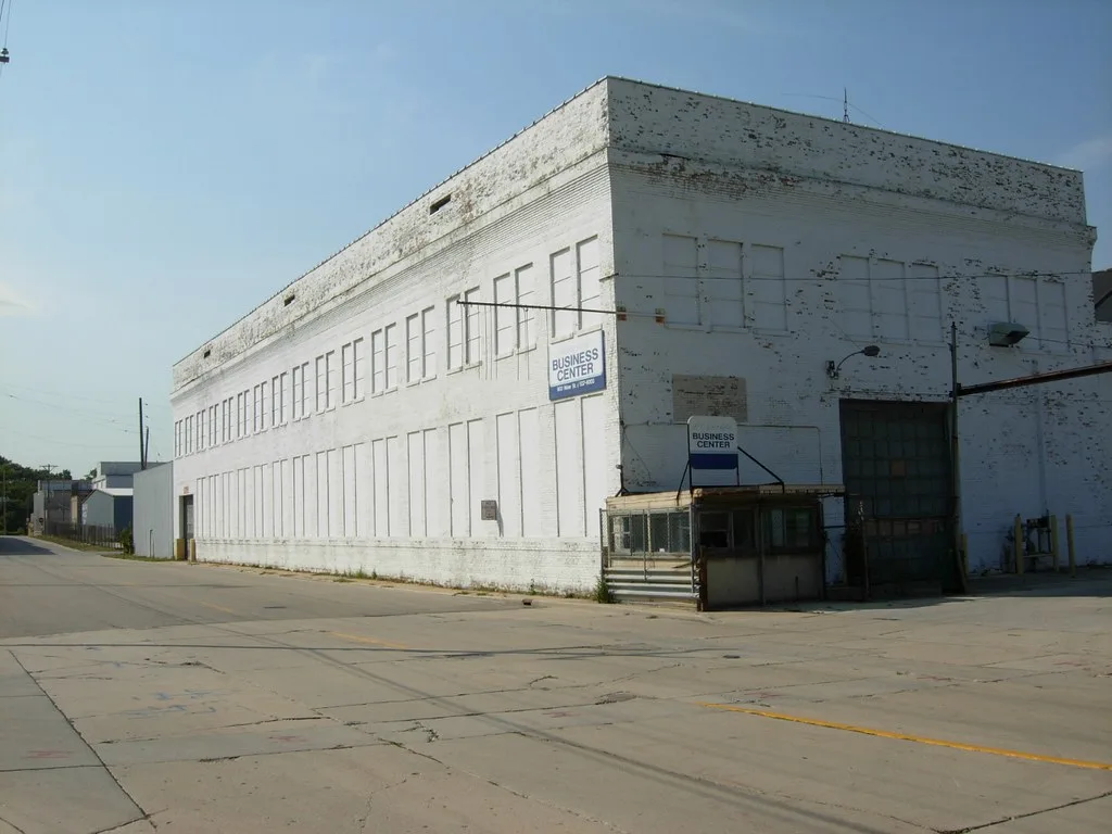 Large white industrial building with sealed windows and a dilapidated guard booth beside its gate.