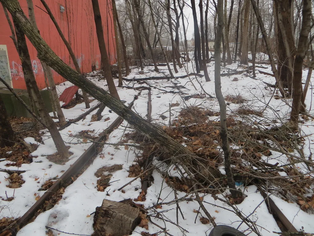 Voies abandonnées sous la neige le long d’un mur rouge, obstruées par des arbres.