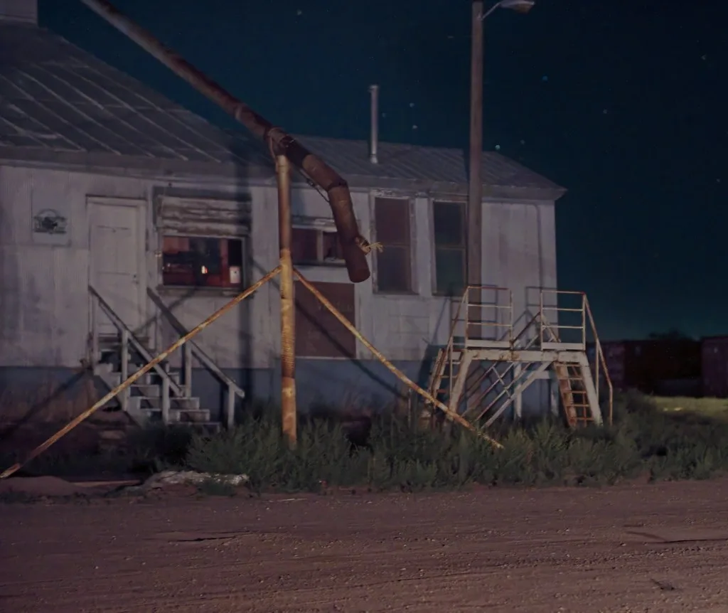 Industrial building at night with boarded windows, rusty stairs and tall weeds.