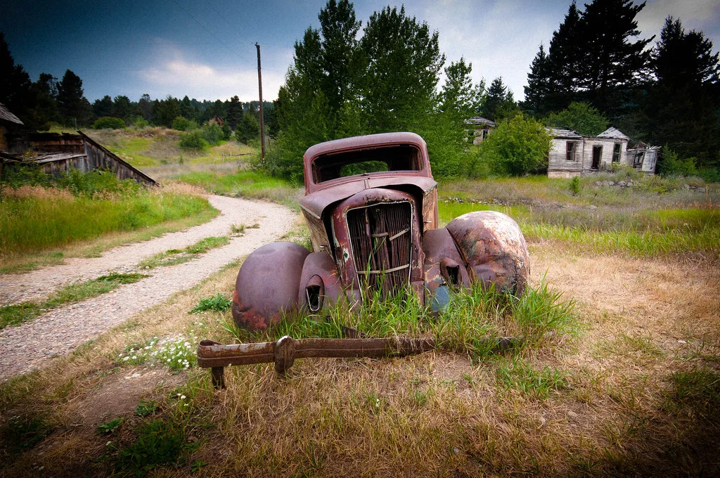 Rusty car wreck in front of derelict wooden houses.
