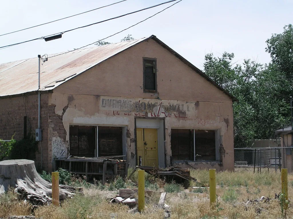 Old shop with a yellow door and boarded windows on overgrown ground.