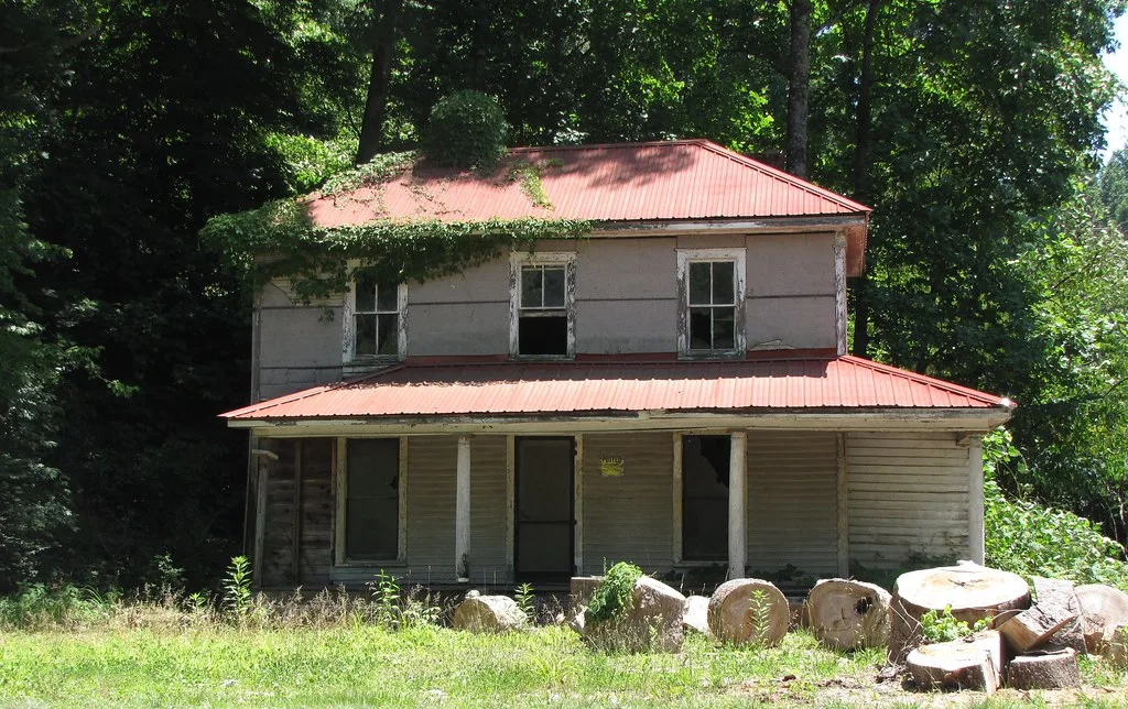 Ancienne maison à deux niveaux au toit rouge et aux fenêtres brisées sous les arbres.