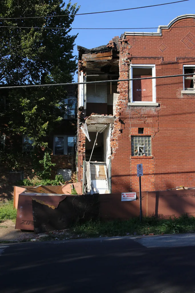 Abandoned red brick building with a collapsed corner exposing two storeys behind a hoarding.
