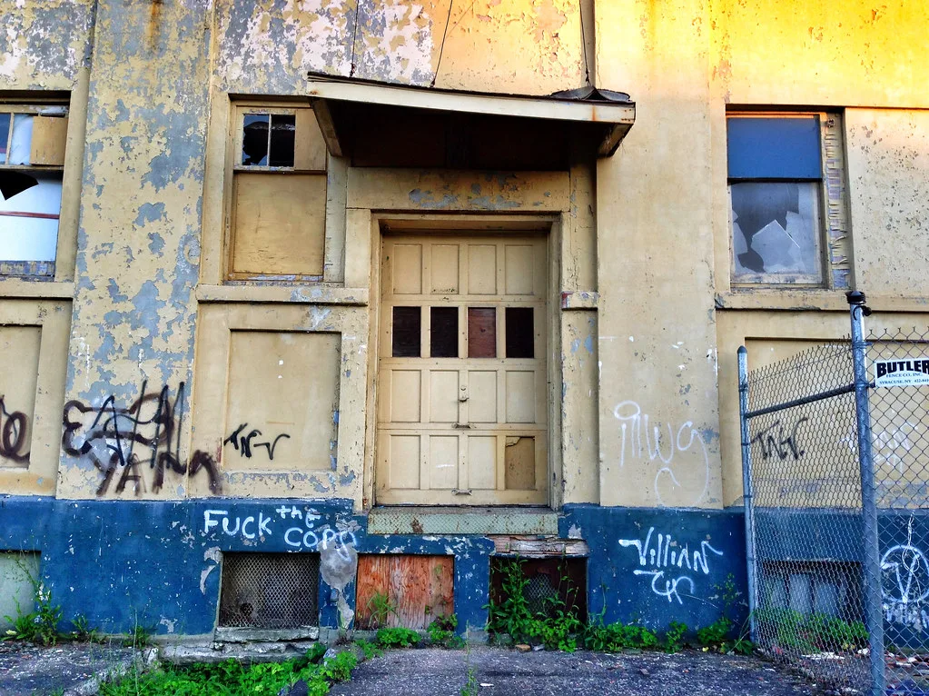 Entrance of an abandoned yellow and blue building with broken windows and peeling graffitied walls behind a fence.