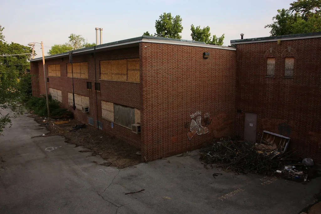 Red brick building with boarded windows overlooking a cracked courtyard littered with debris.