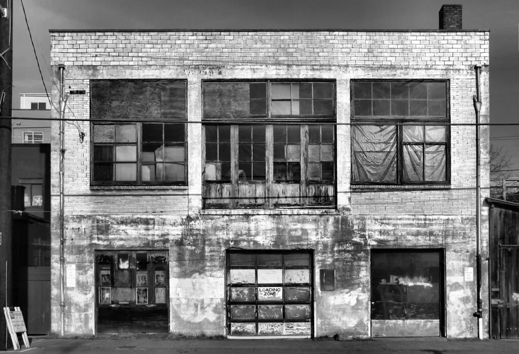 Facade of an old workshop with damaged large windows, in black and white.