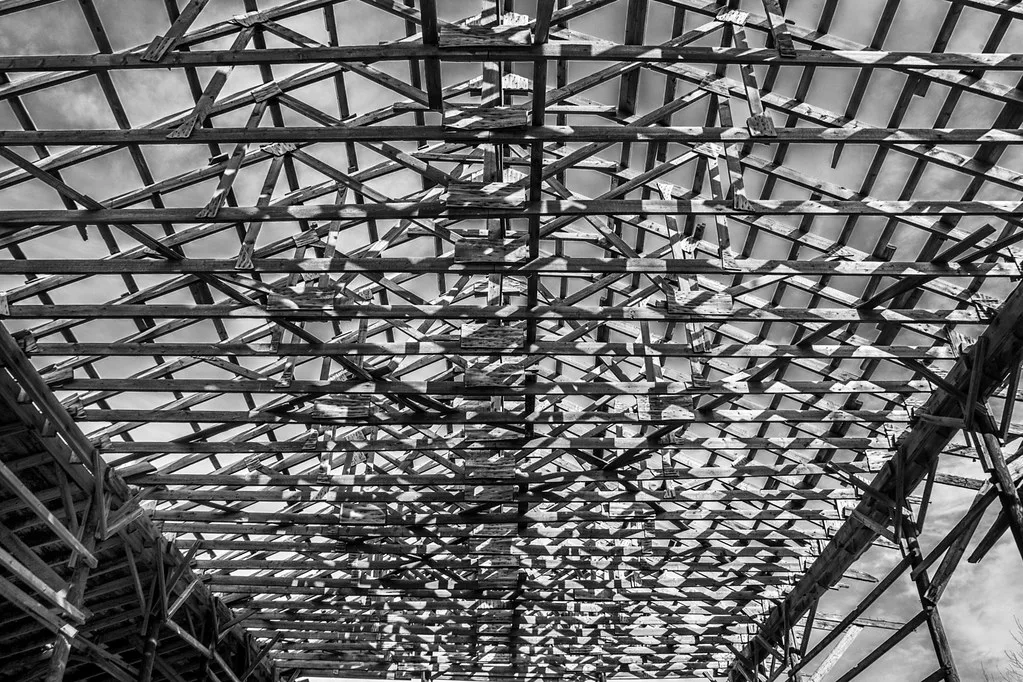 Wooden roof framing of an abandoned building without covering, with separated timbers silhouetted against the sky in black and white.